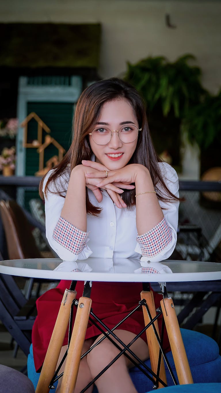 Charming Asian woman with glasses, smiling confidently while seated at a table indoors.
